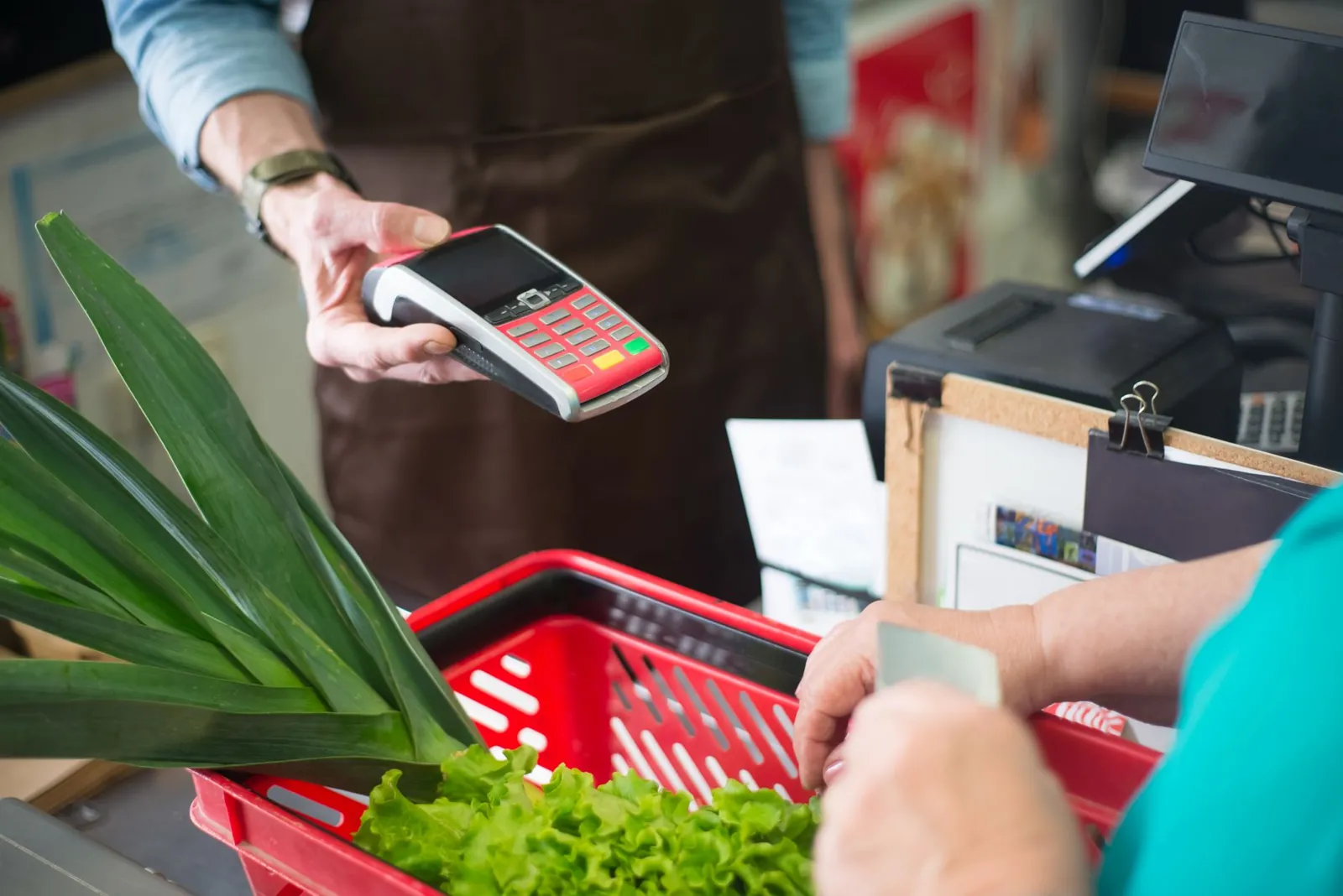 Paiement par carte à la caisse d'un commerce, avec un panier rouge contenant des poireaux et de la laitue