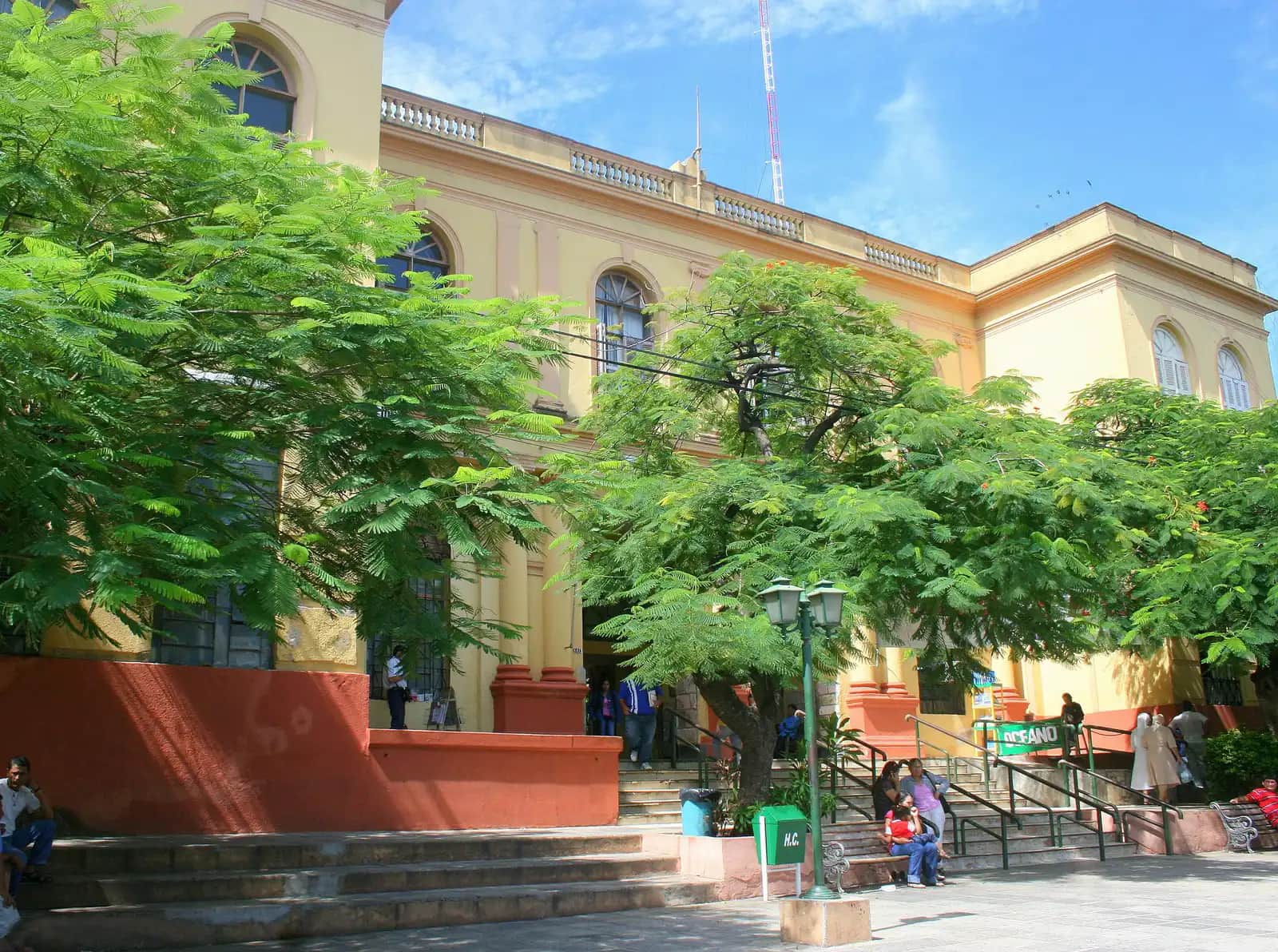 Façade jaune et ocre de l'Hospital de Clínicas à Asunción, derrière des flamboyants, avec des patients assis sur les marches