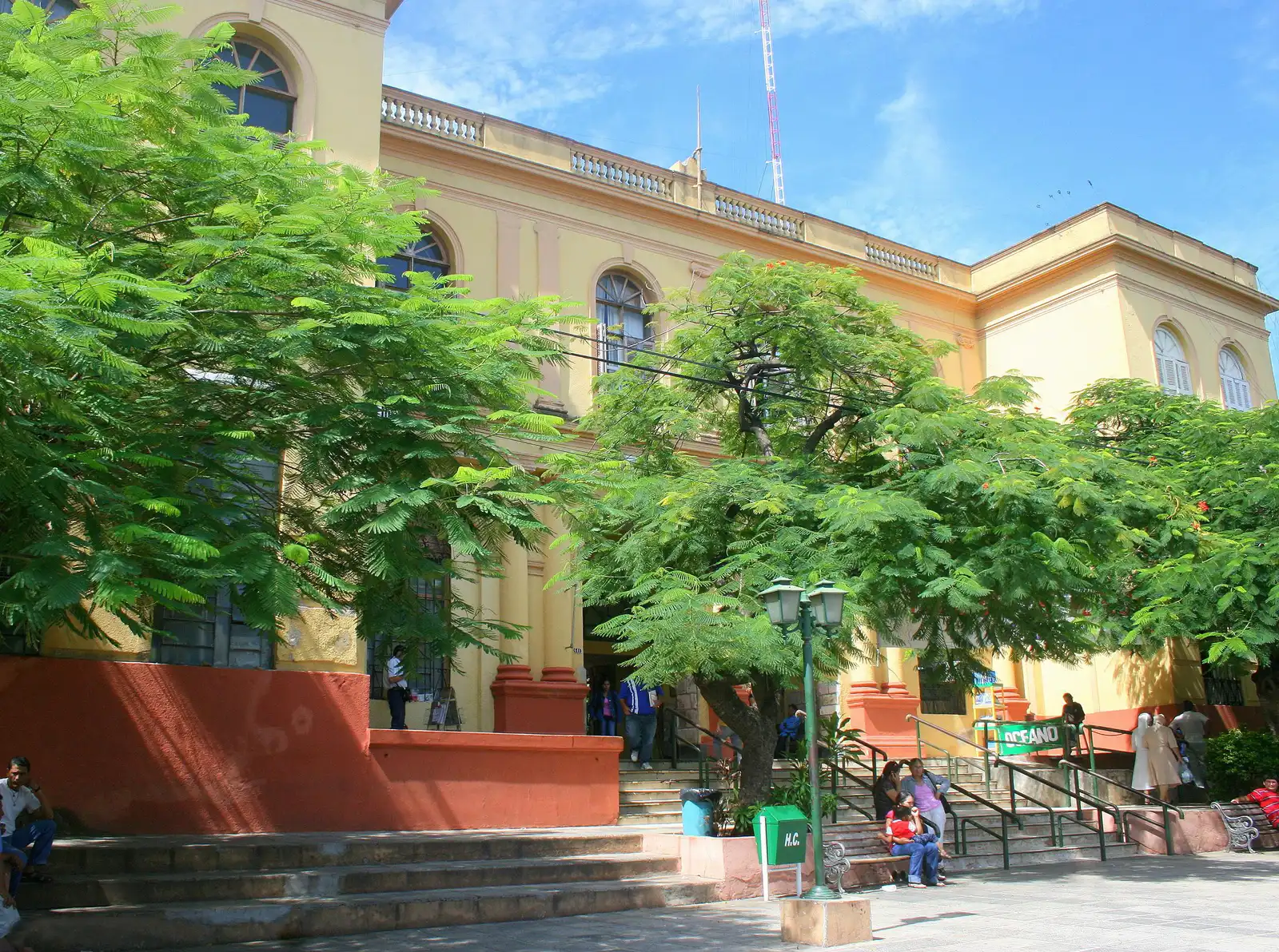Façade jaune et ocre de l'Hospital de Clínicas à Asunción, derrière des flamboyants, avec des patients assis sur les marches