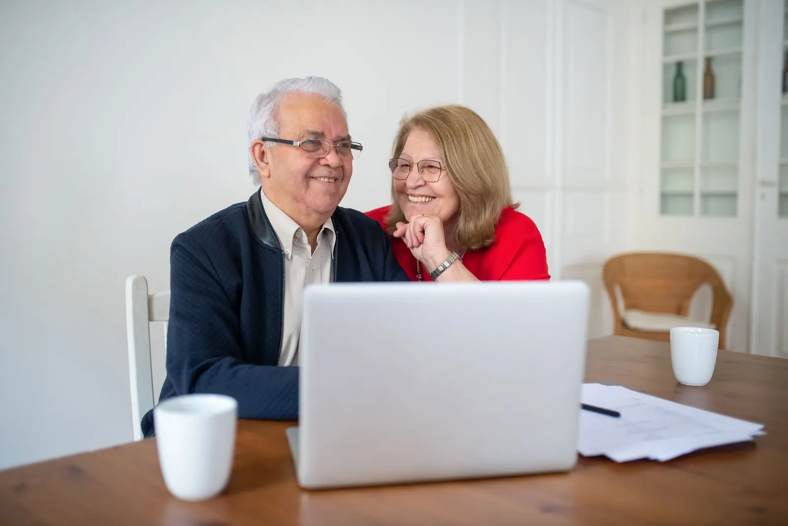 Couple de retraités souriants assis à une table devant un ordinateur portable, avec des documents posés à côté