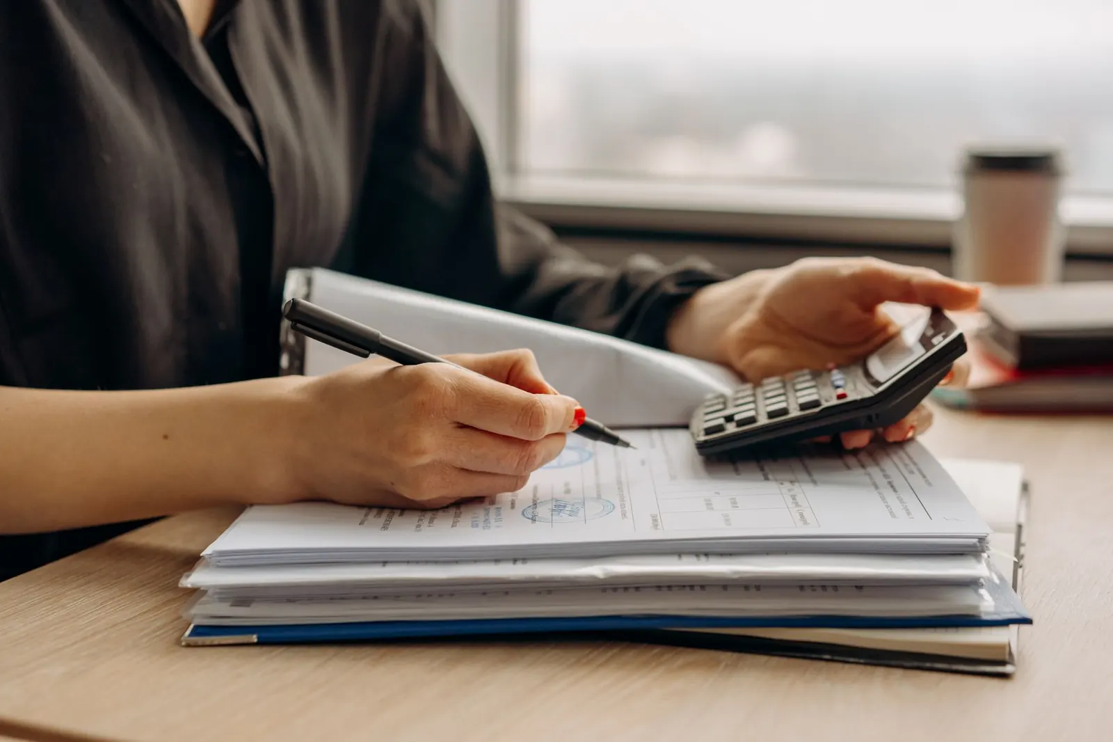 Mains sur une pile de documents avec stylo et calculatrice, sur un bureau près d'une fenêtre