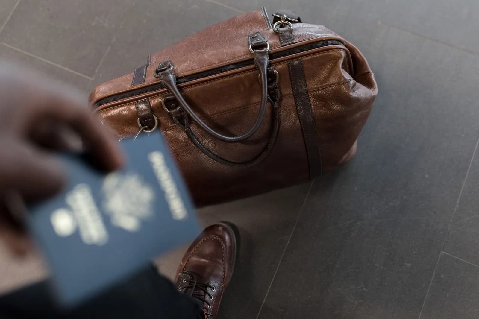 Une main tient un passeport à côté d'un sac de voyage en cuir posé sur un sol carrelé