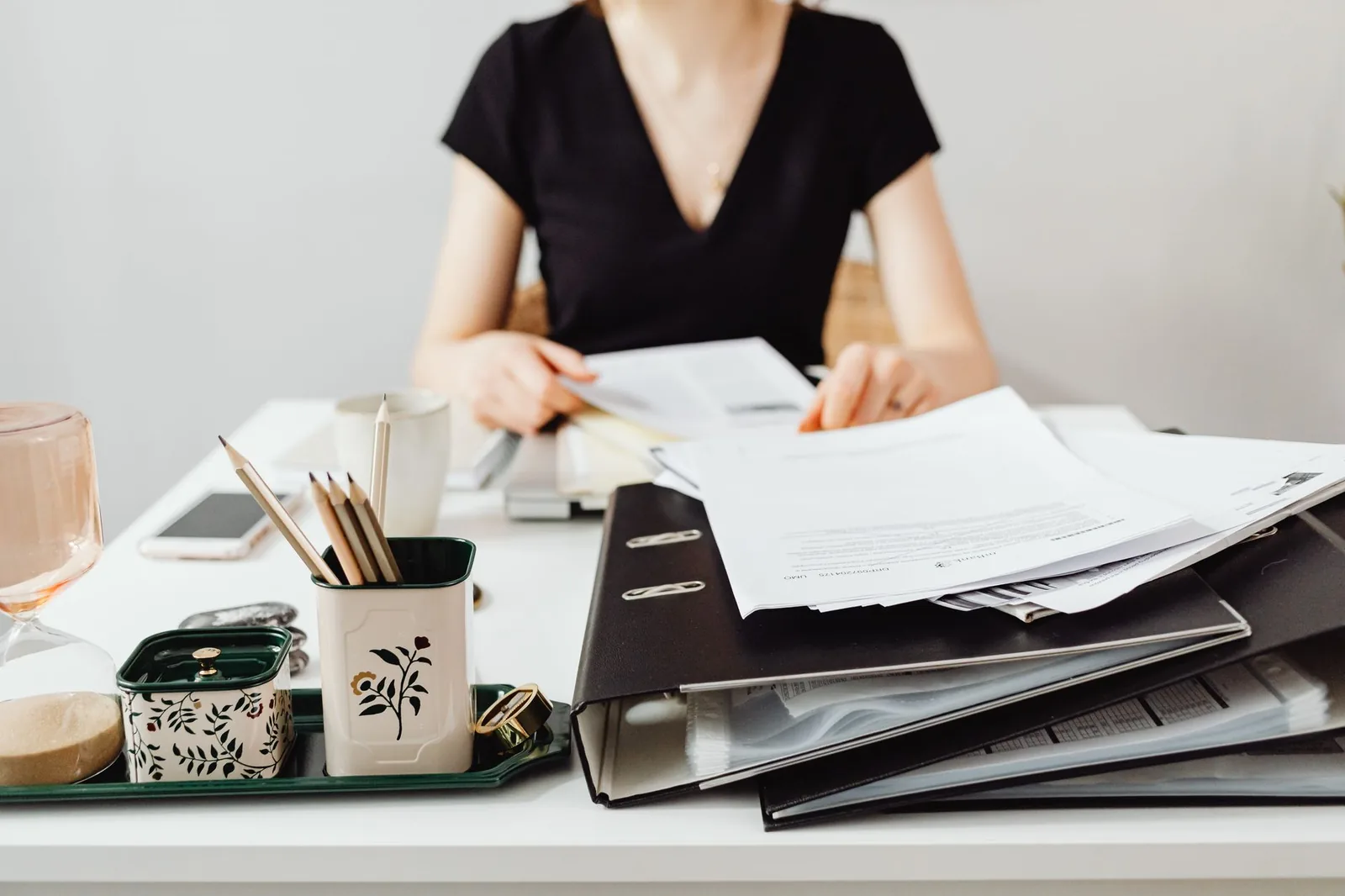 Bureau avec classeurs noirs et feuilles de documents, une personne dont le visage n'est pas visible consulte un dossier