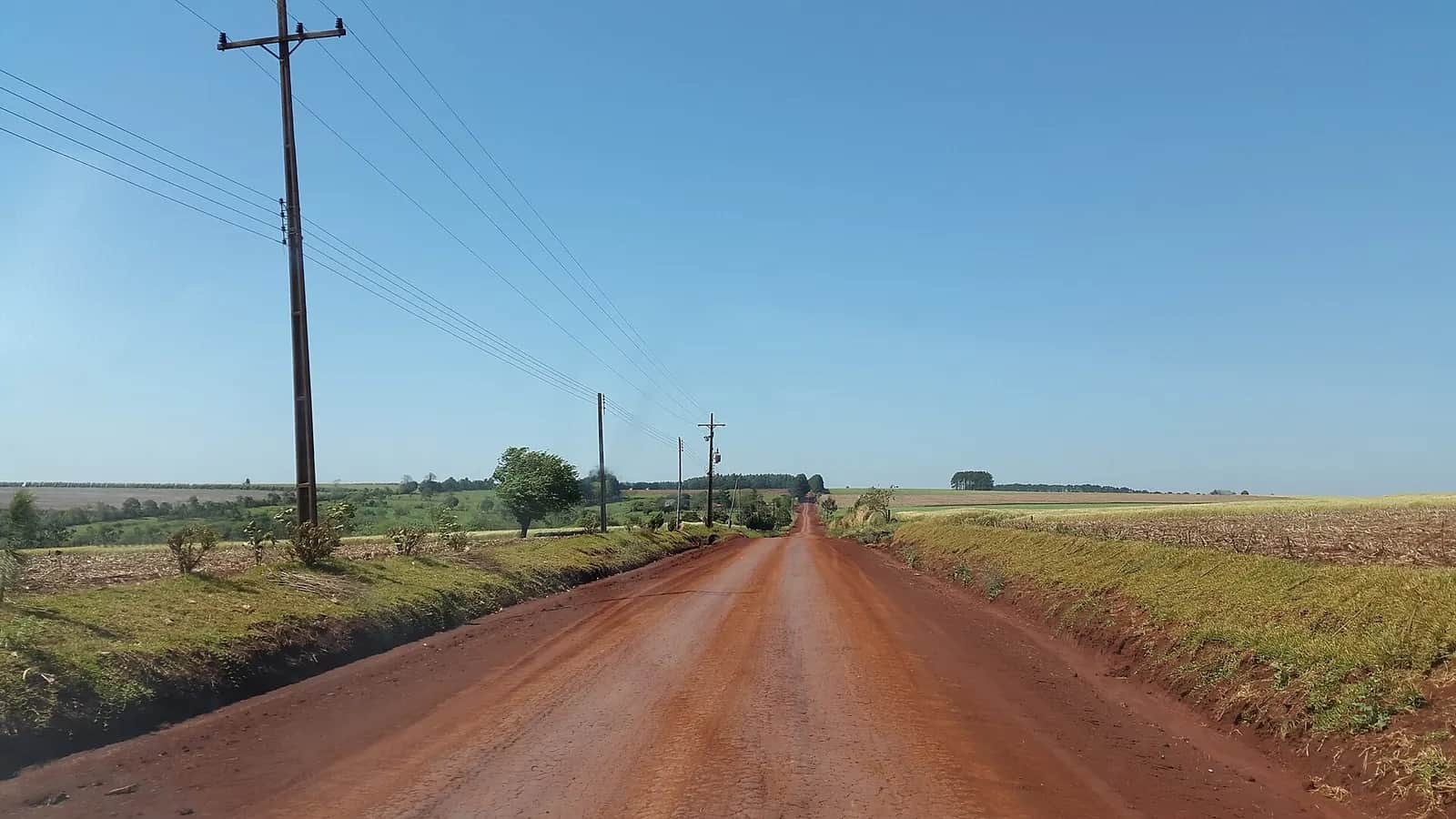 Route de terre rouge bordée de champs cultivés et de poteaux électriques, dans la campagne de Pirapó, département d'Itapúa, Paraguay
