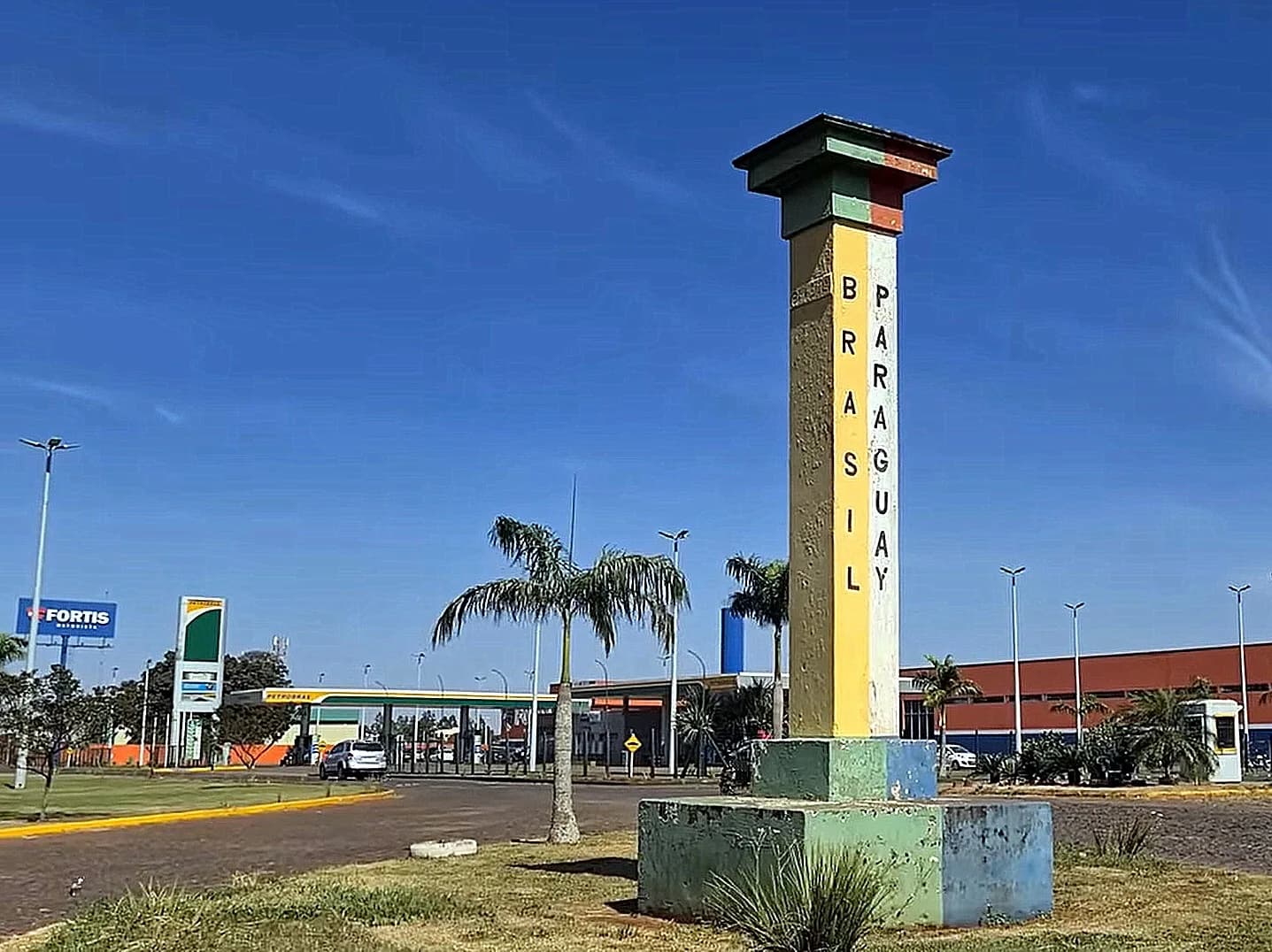 Borne jaune, verte et bleue marquée Brasil et Paraguay à la frontière entre Ponta Porã (Brésil) et Pedro Juan Caballero (Paraguay), avec une station-service et des palmiers