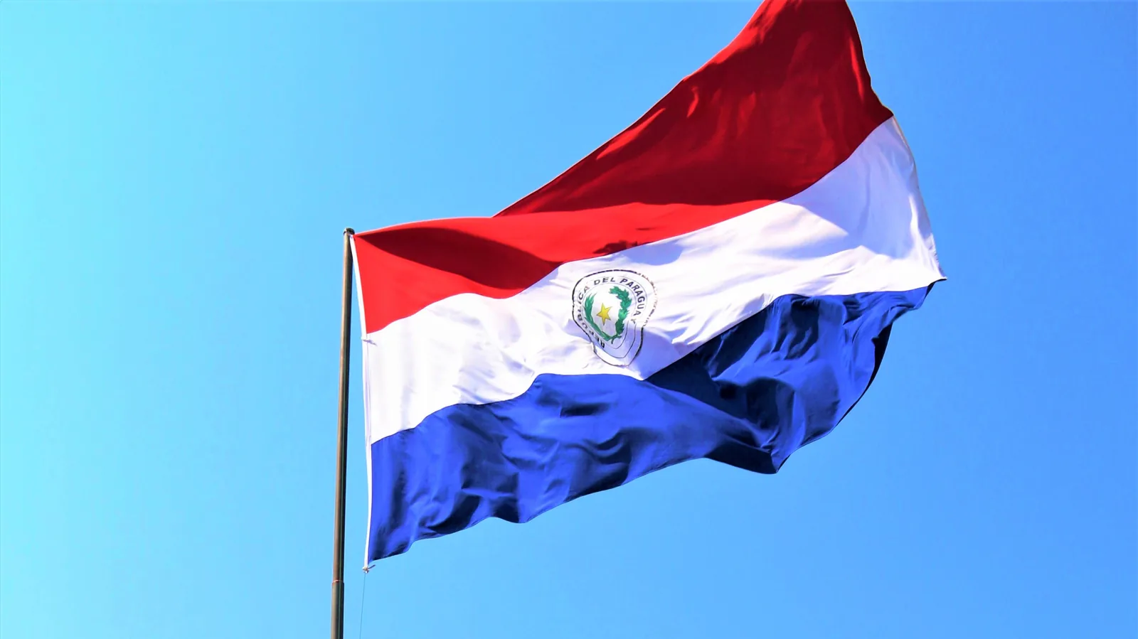 Drapeau tricolore du Paraguay (rouge, blanc, bleu) flottant sur un mât devant un ciel bleu