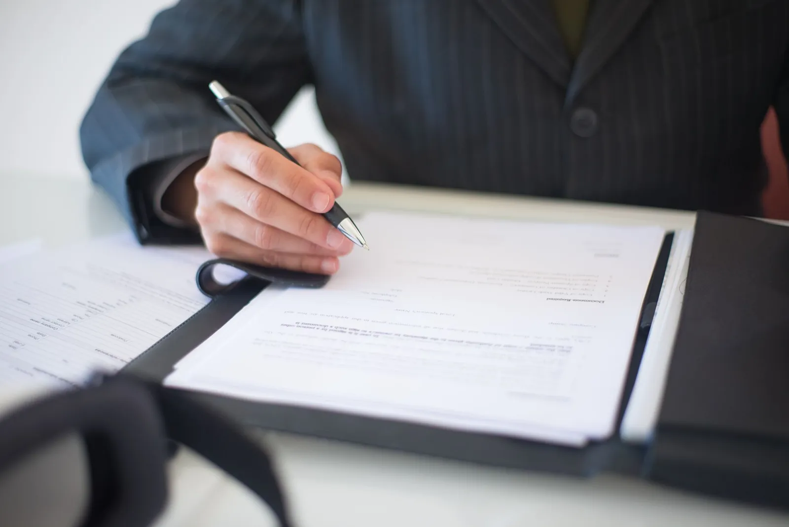 Main d'une personne en costume signant des documents avec un stylo, dossier ouvert sur un bureau